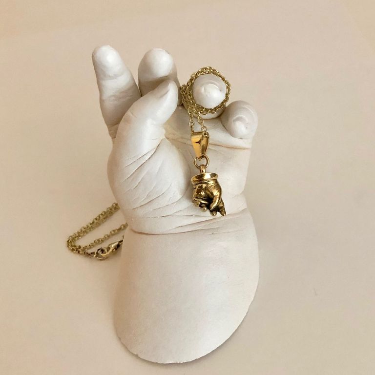 A life size plaster cast of a baby's hand with a miniature version in yellow gold on a chain draped over it