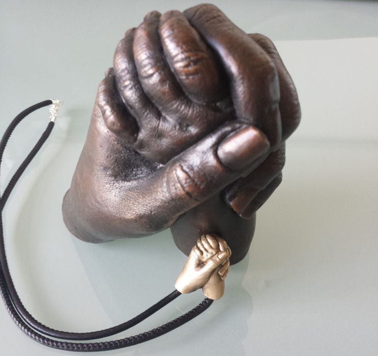 A life size real bronze cast of dad holding his baby daughter's hand with a miniature bronze version set on a leather necklace 