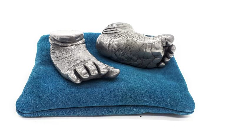 Two aluminium resin baby foot casts placed on a teal coloured cushion, one foot angled on its side to see the detail of the sole of the foot